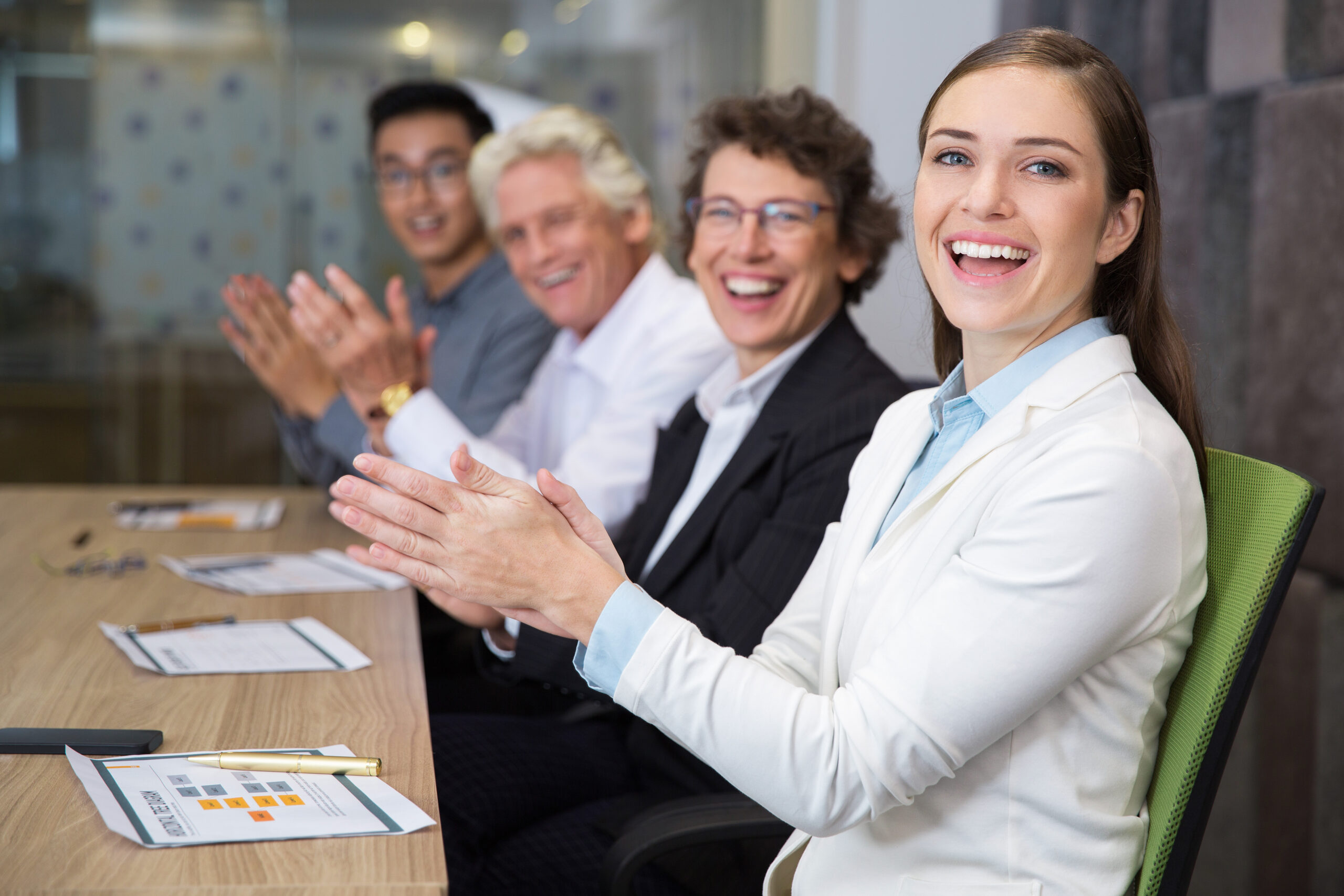 Cheerful multiethnic business people sitting at meeting in boardroom, clapping, looking at camera and laughing. Young woman sitting in front